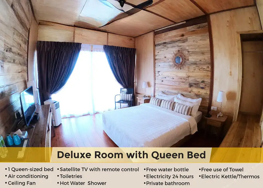 A deluxe bedroom at Dreamy Wave Dwelling Resort featuring a queen bed facing panoramic glass doors framing the seaviews, warm wooden plank walls with varnished timber flooring, and air con.